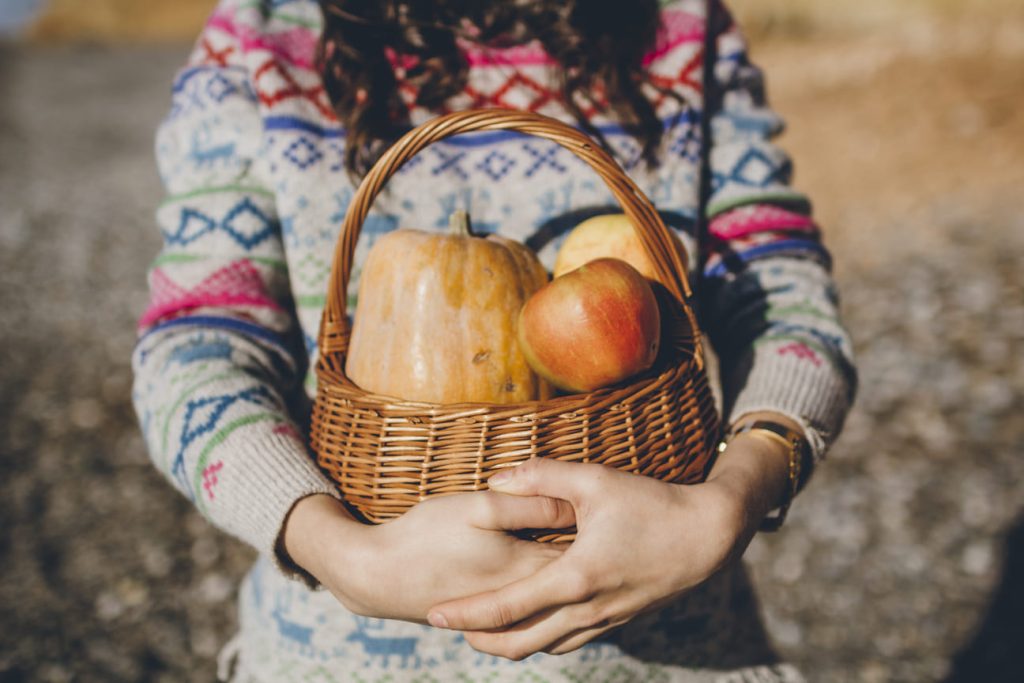 Persona sosteniendo una cesta con calabazas y manzanas.